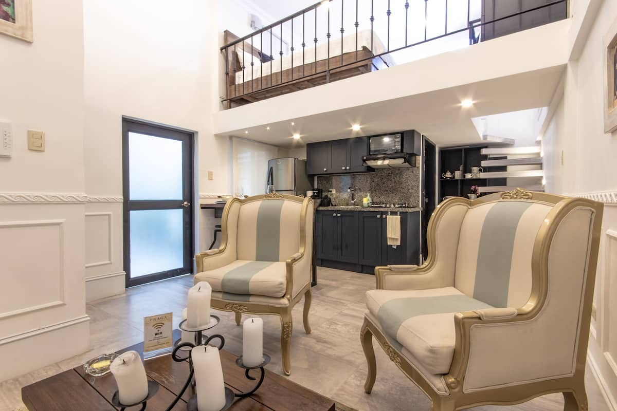 A spacious living area with two ornate armchairs, a coffee table with candles, and a view of the kitchen and balcony.