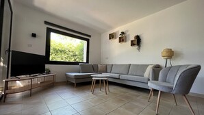 Uma sala de estar moderna com um sofá cinzento, uma mesinha redonda, uma TV sobre um suporte e uma janela com vista para o verde.