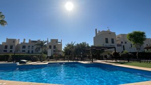 Uma piscina com superfície de água azul cristalina, rodeada por um deck revestido de azulejos. Ao fundo, edifícios modernos com várias janelas e varandas.