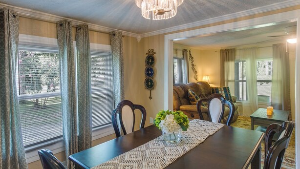 A dining area with a dark wooden table, white placemat, and a vase with flowers. There are chairs around the table, and a window with blinds and curtains. - Waycross Vacation Rental w/ Private Backyard! (Waycross)