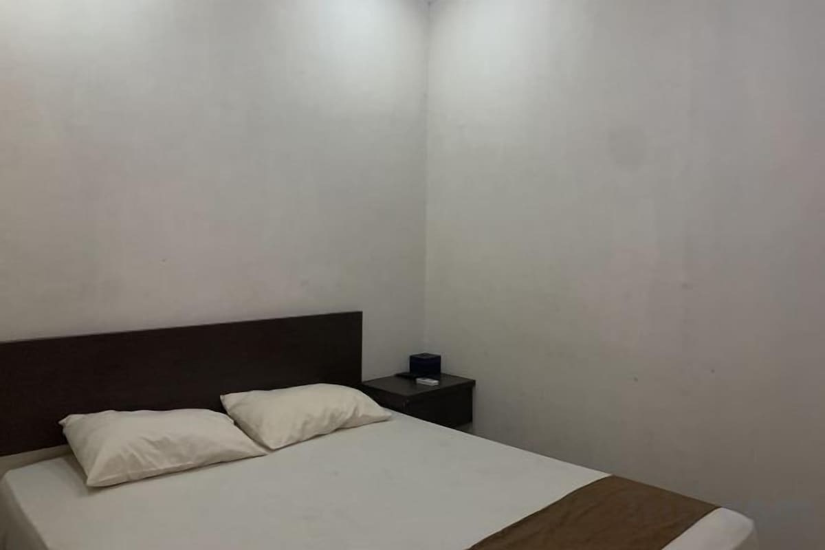 A single bed with a wooden headboard and two pillows, a bedside table with a lamp, and a plain wall.
