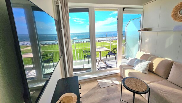 A modern living room with a large window offering a view of the sea and a balcony with outdoor furniture. - One-room apartment Kaiserhof 311 Norderney with sea view directly on the beach (Norderney)