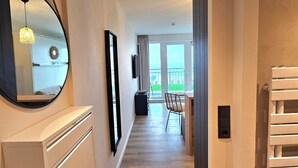 A hotel hallway with a wooden floor, a mirror, a ceiling light, a chair, and a view of a balcony. - One-room apartment Kaiserhof 311 Norderney with sea view directly on the beach (Norderney)