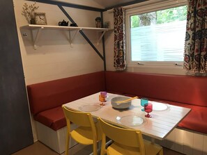 Een eethoek met een witte tafel, gele stoelen en rode bankjes. Daarboven staan planken met een klok en decoratieve objecten.