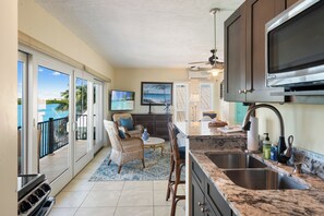 A kitchen with a large island, a dining area with wicker chairs, a view of the ocean through sliding glass doors, and a ceiling fan.