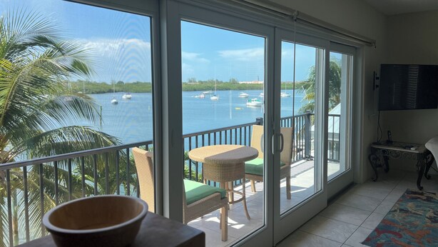 Ein Zimmer mit großen Fenstern mit Blick auf ein Gewässer und Boote, ein Balkon mit Korbmöbeln und einem Flachbildfernseher.