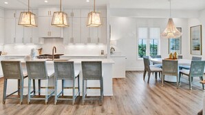 Una cocina moderna con armarios blancos, una isla central con taburetes de bar, iluminación colgante y un comedor con una mesa redonda y sillas.