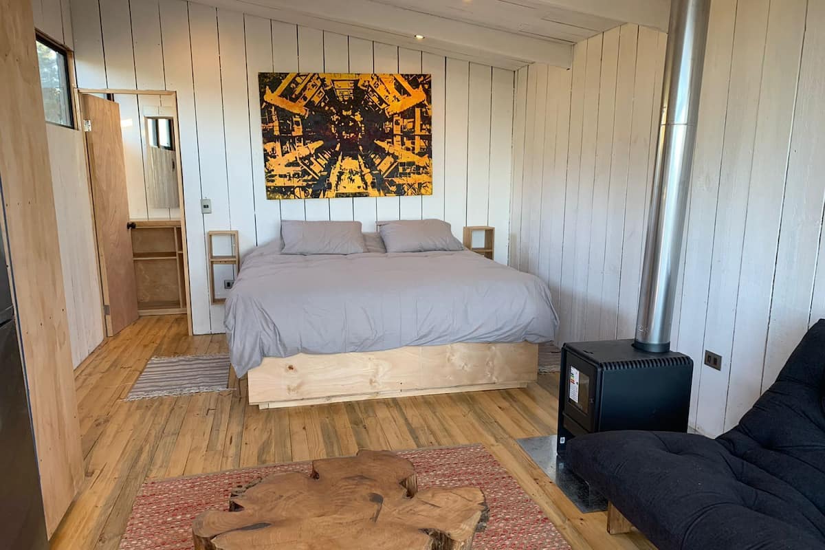 Un dormitorio con cama, piso de madera, un cuadro grande en la pared, estufa de leña y sofá oscuro.