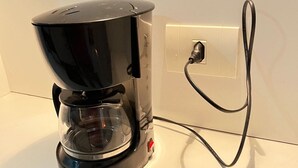 A black coffee maker with a transparent water reservoir, plugged into a white wall socket. - Stúdio Praia Ipanema (Rio de Janeiro)
