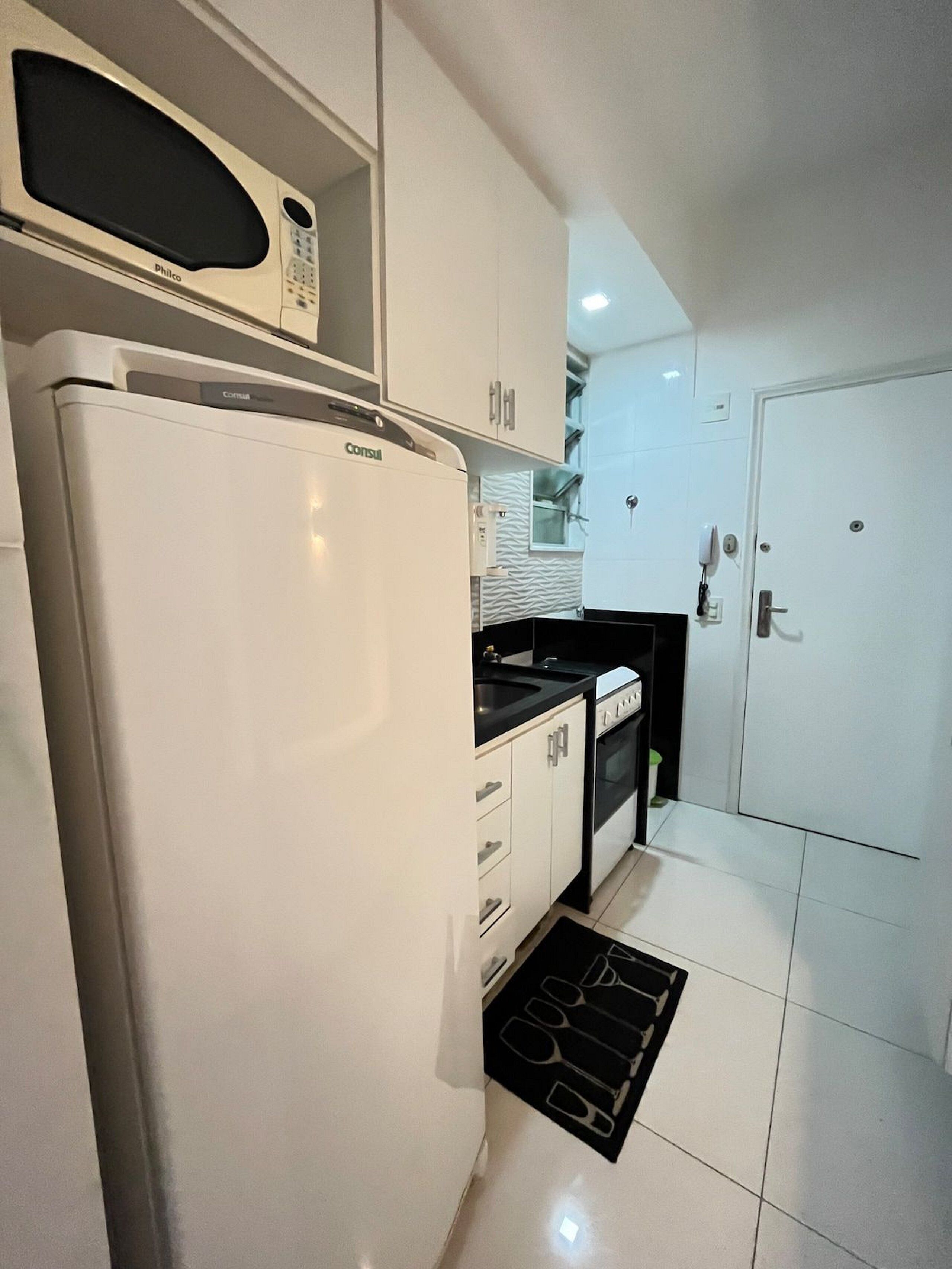 A compact kitchen with a white refrigerator, a microwave, a stove, and a sink. There is a patterned rug on the floor and a door to the right.