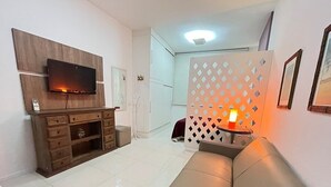 A living room with a flat-screen TV mounted on a wooden panel, a leather sofa, a small table with a candle, and a decorative partition with diamond cut-outs. - Stúdio Praia Ipanema (Rio de Janeiro)