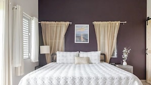 Un dormitorio con una cama grande, dos mesitas de noche, una mesita auxiliar, un cuadro en la pared y una ventana con cortinas.