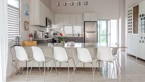 Una cocina moderna con una isla central, taburetes blancos de bar, electrodomésticos de acero inoxidable y un amplio ventanal que proporciona luz natural.