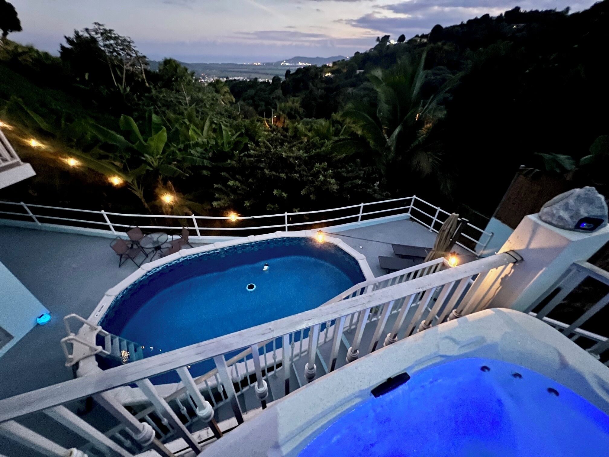 A pool area with a hot tub, surrounded by a white railing, and overlooking a lush landscape with trees and distant hills.