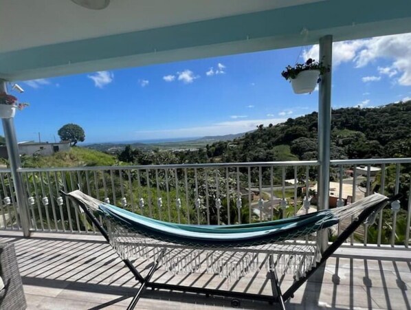 Un hamac sur un balcon, avec vue sur un paysage verdoyant et des bâtiments éloignés.