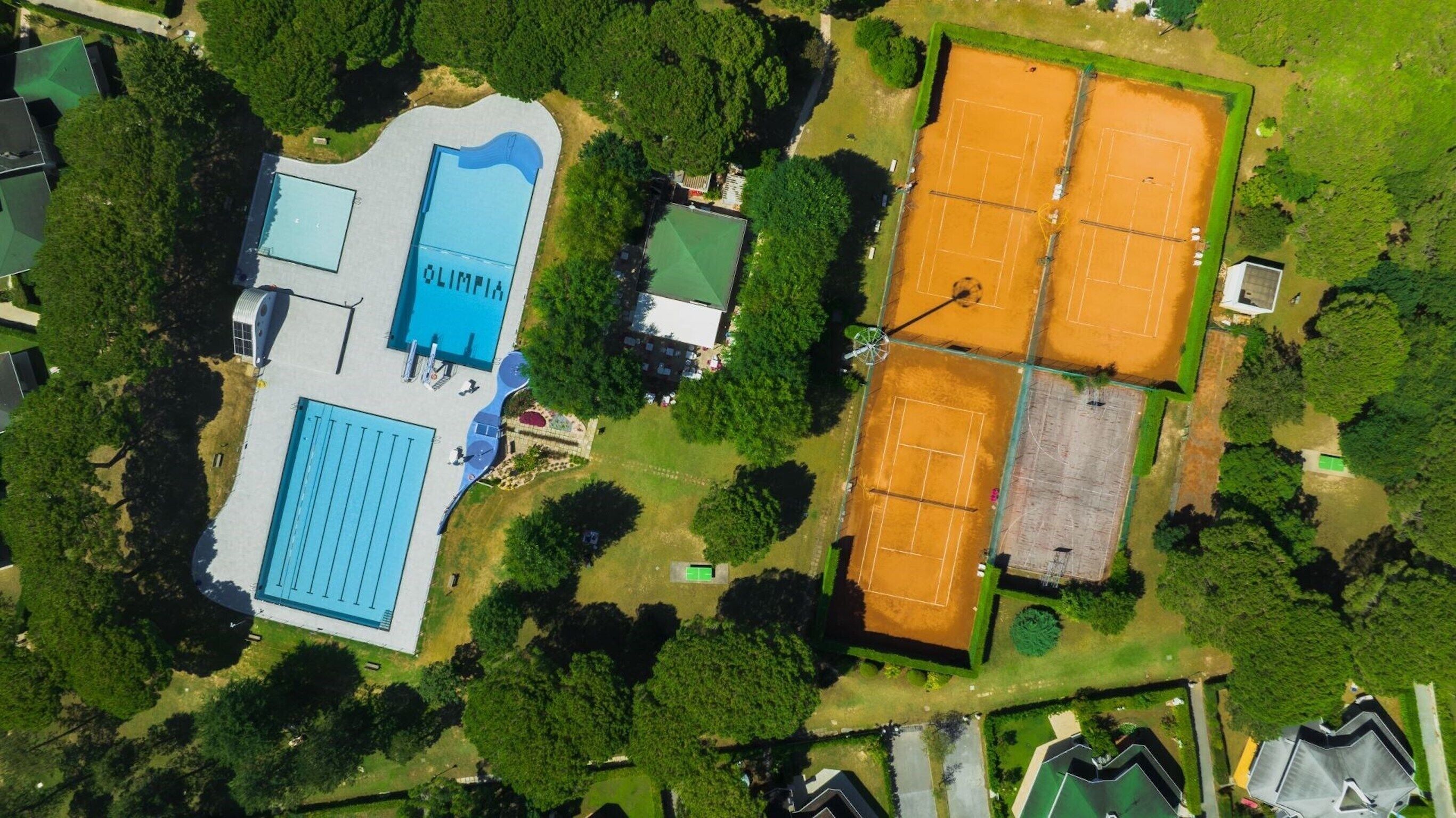 An aerial view of a sports complex with multiple tennis courts, a swimming pool, and surrounding green areas.
