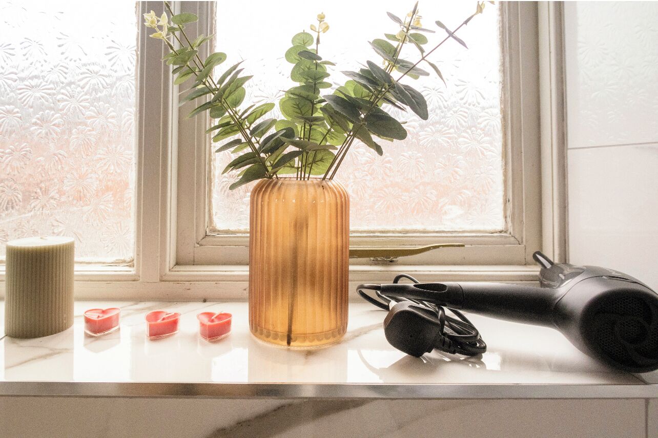 A window with a plant in a vase, a hairdryer, and three candles on a windowsill.