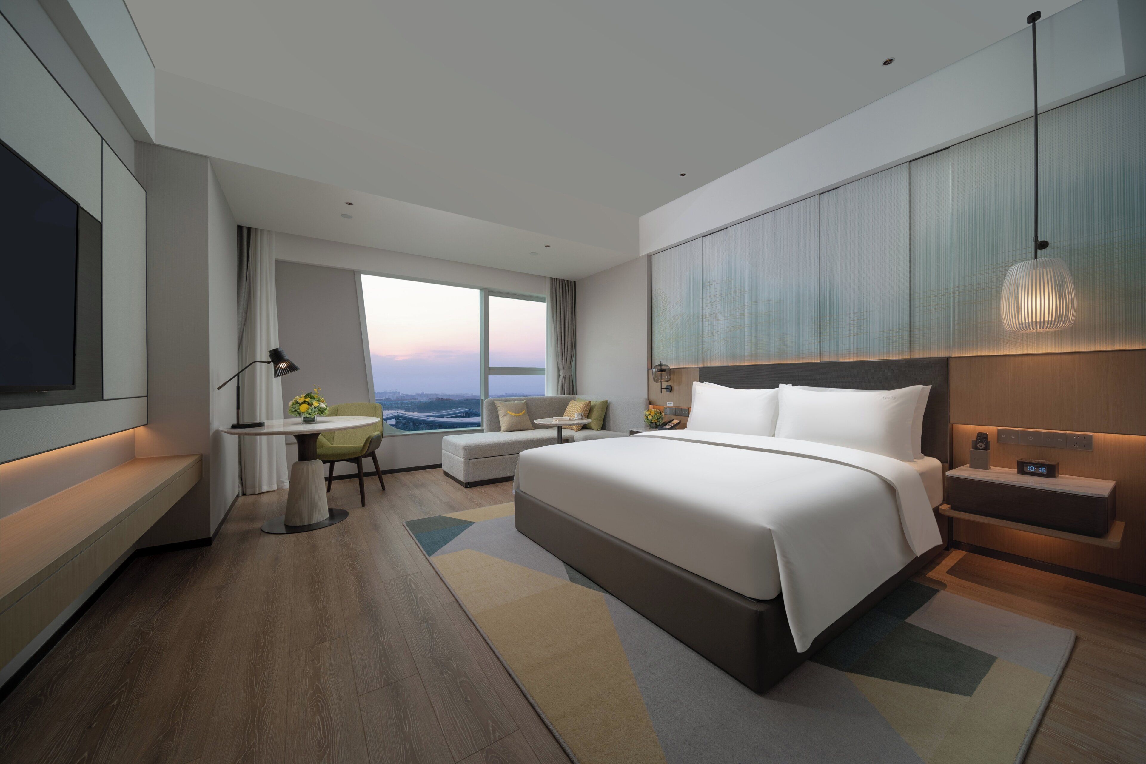 A modern hotel room with a large bed, a desk with a chair, a sofa, and a view of the cityscape.