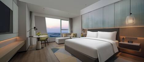 A modern hotel room with a large bed, a desk with a chair, a sofa, and a view of the cityscape.