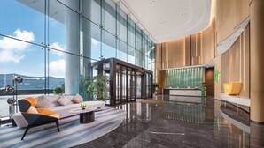 Um hall amplo com uma recepção moderna, grandes janelas de vidro e uma área de estar com sofá e poltrona.