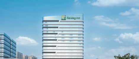 A modern Holiday Inn hotel with a glass facade, surrounded by greenery and other buildings.