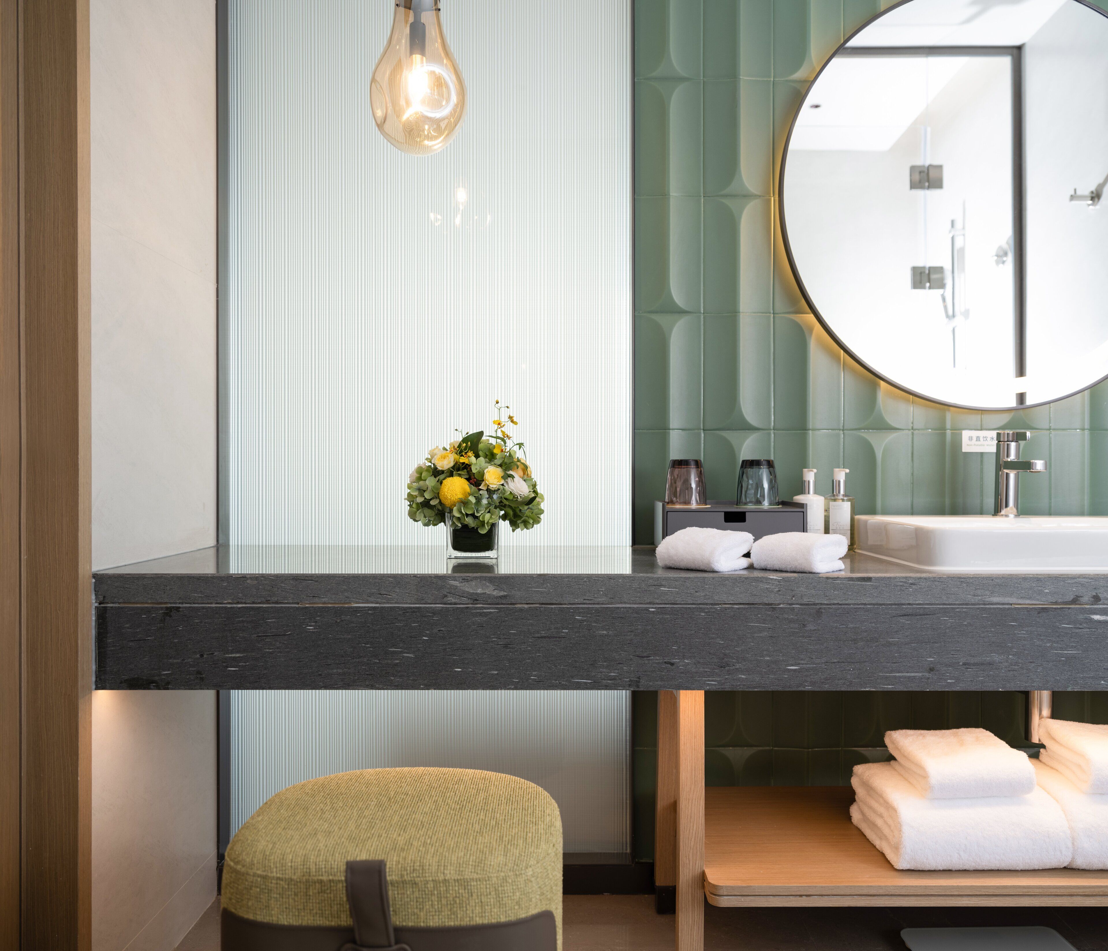 Un bagno moderno con un piano di lavoro in marmo, uno specchio rotondo, un piccolo vaso con fiori e asciugamani piegati con cura.