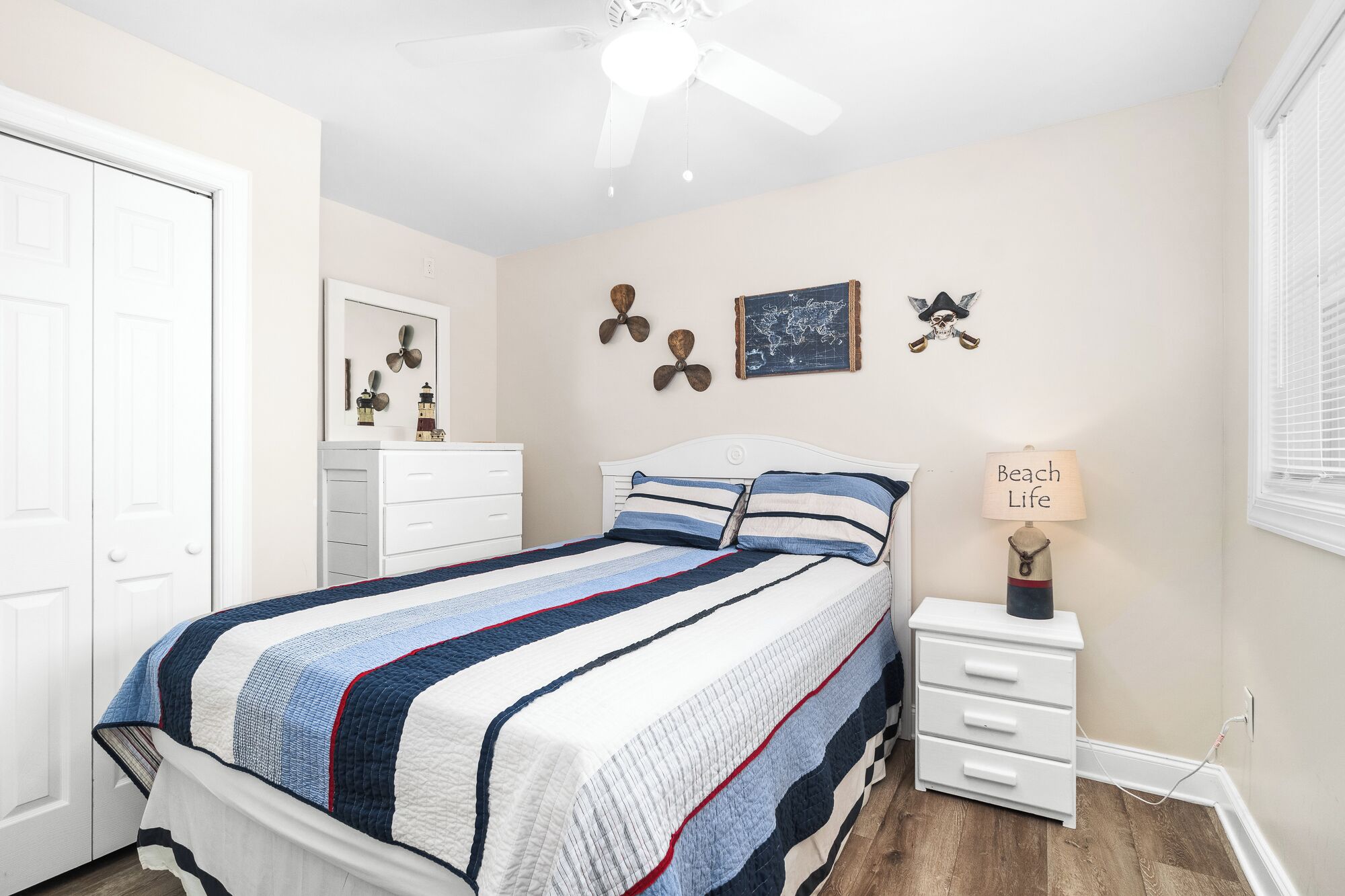 Une chambre à coucher comprenant un lit, une commode, une table de chevet, un ventilateur de plafond et des éléments de décoration murale, notamment une carte du monde et un crâne.