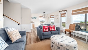 A living room with a grey sofa, a round ottoman, and a dining area with a table and chairs. There is a staircase on the left and a view of the ocean through the windows. - Oceanfront Updated 5BR Duplex. Dog friendly! (Emerald Isle)