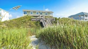 A coastal house with a staircase leading to a deck, surrounded by tall grass and a clear blue sky. - Oceanfront Updated 5BR Duplex. Dog friendly! (Emerald Isle)