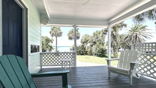 A covered porch with wooden flooring, two green Adirondack chairs, a white chair, and a white lattice fence. - 1 Minute Walk to Ocean-Charming Downtown Beachside Home with Mother-In-Law Suite (Cocoa Beach)
