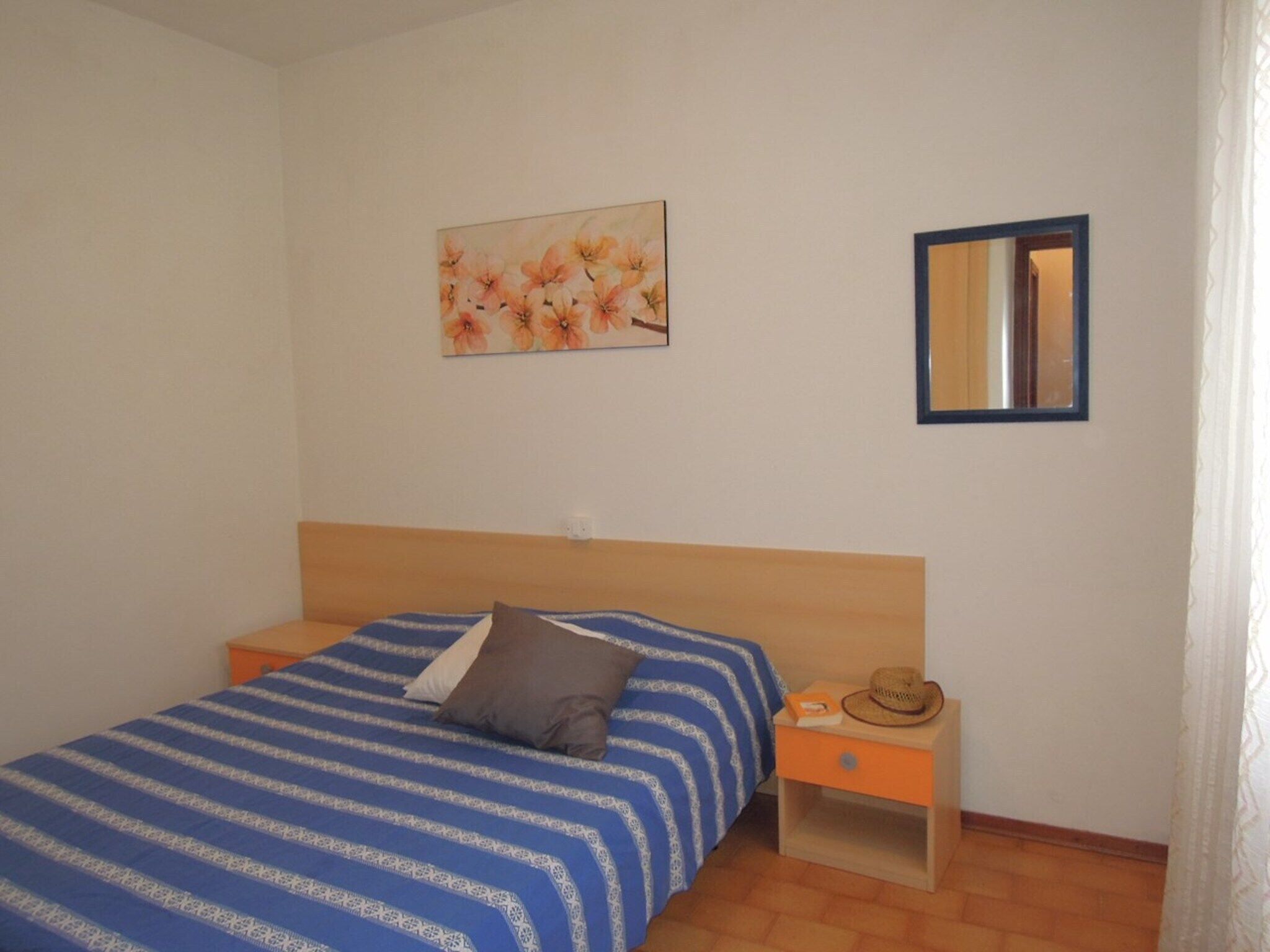 A bedroom with a bed, a nightstand, a mirror, and a floral painting on the wall.