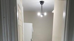 Un pasillo estrecho con una puerta blanca a la izquierda, un electrodoméstico blanco a la derecha y una lámpara en el techo.