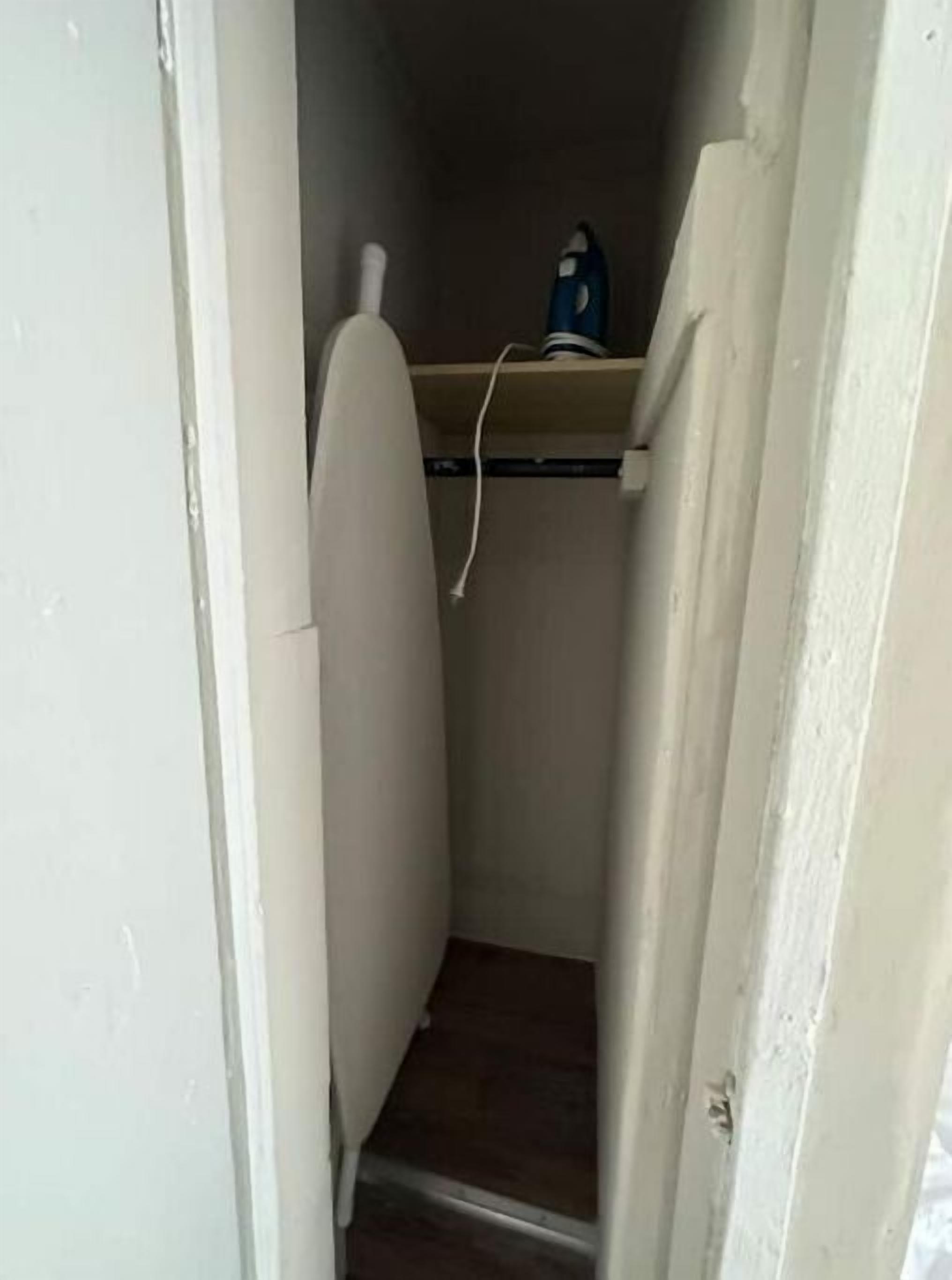 Un pasillo estrecho con una puerta blanca a la izquierda y una habitación pequeña con una pared blanca y un objeto azul sobre una estantería.