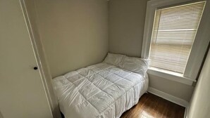 Una habitación pequeña con cama individual, suelo de madera, ventana con persianas y una puerta.