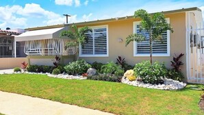 A single-story house with a beige exterior, white window shutters, and a small front yard with various plants and a pathway. - My Escape Dreamy Luxury Home in Bayamón, P.R. (DAVILA BAYAMON PR)