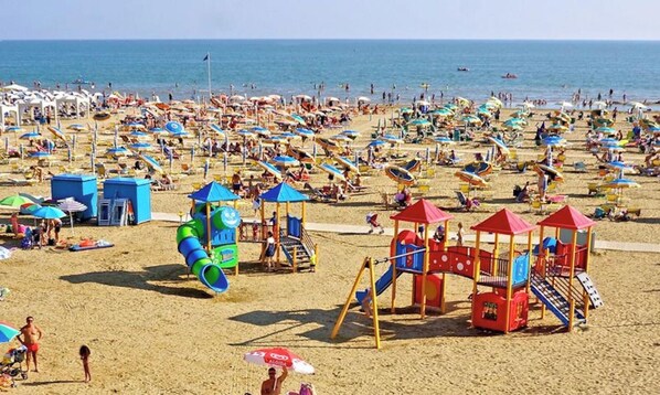 A crowded beach with numerous umbrellas, a children's play area, and a clear view of the sea. - Nice and Cozy Studio With Pools (San Michele al Tagliamento)