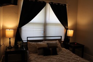 Une chambre à coucher avec un lit, deux tables de chevet avec des lampes, une fenêtre avec des stores et un rideau.