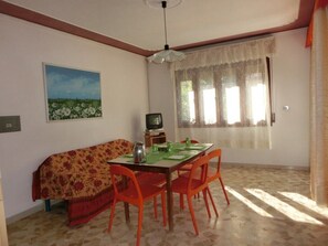 Un coin repas comprenant une table, des chaises, un canapé, un tableau, un téléviseur et une fenêtre ornée de rideaux en dentelle.