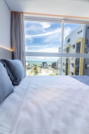 Une chambre d’hôtel avec une grande fenêtre, un lit et une vue sur la plage et les bâtiments.
