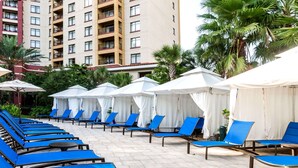 Un espace aménagé au bord de la piscine, avec des chaises longues bleues et des parasols blancs, entouré de palmiers et dominé, au loin, par un immeuble de grande hauteur.