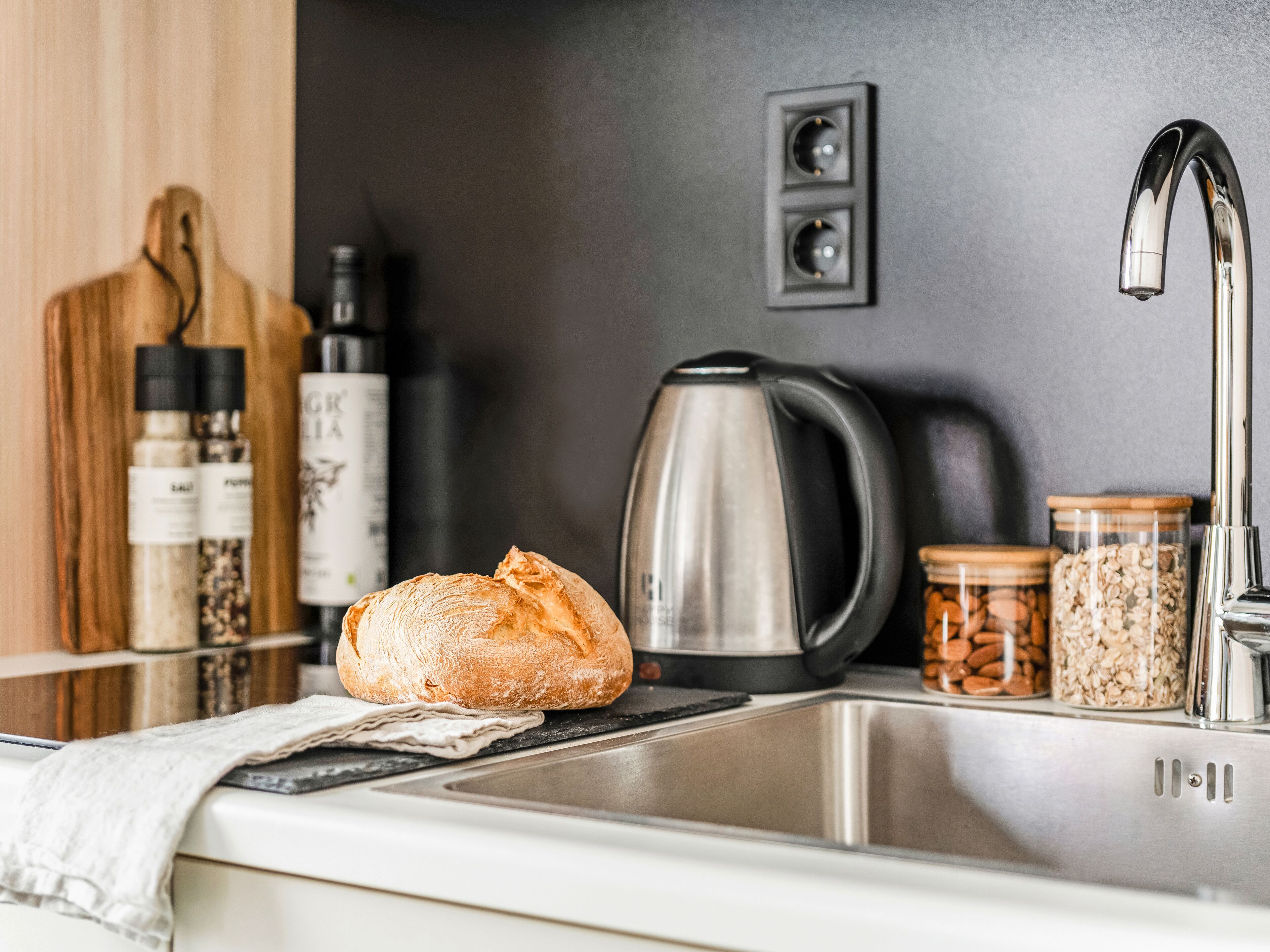A kitchen with a stainless steel sink, a kettle, jars of nuts and oats, and a loaf of bread on the counter.