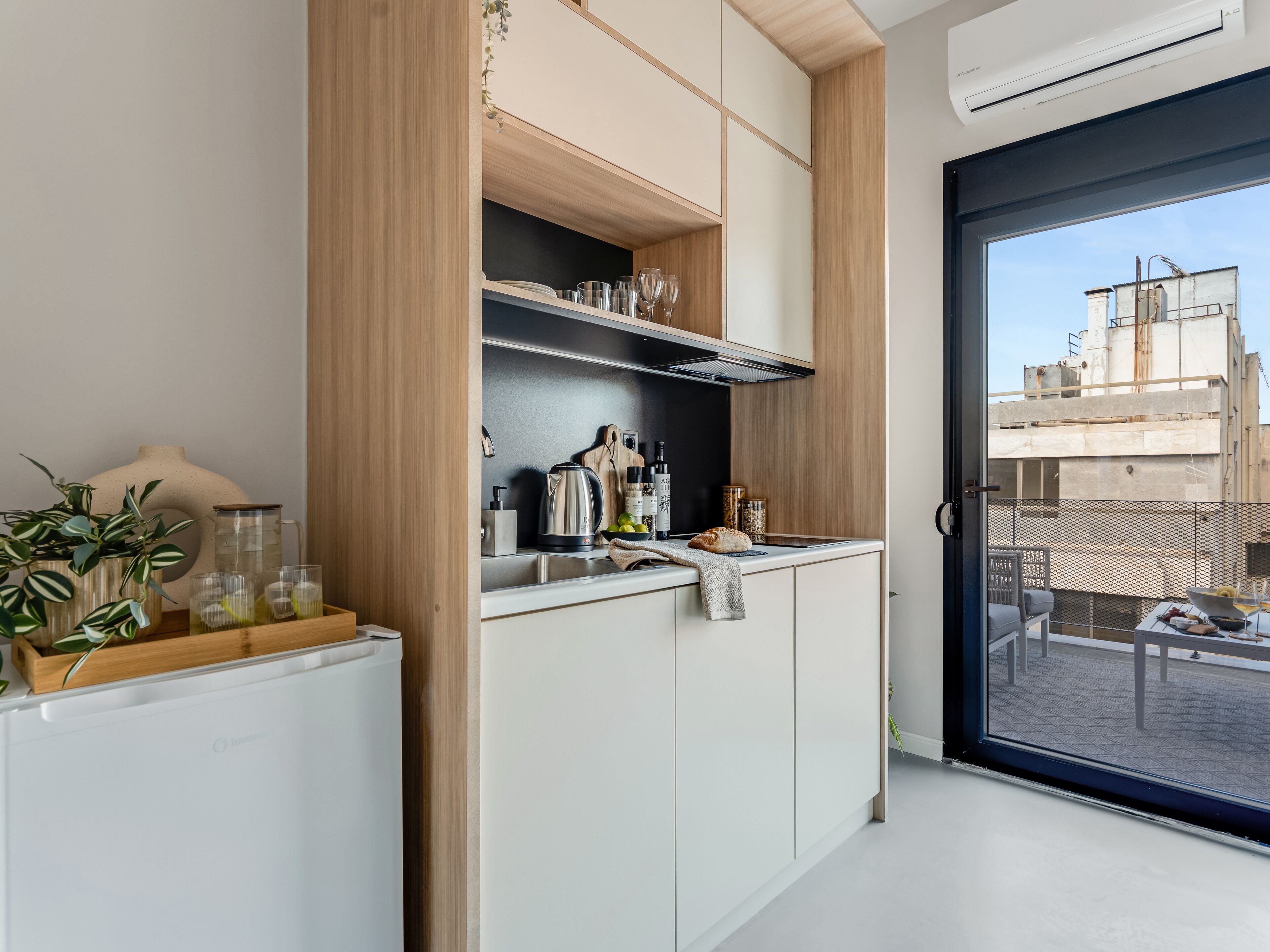A modern kitchen with white cabinets, a stainless steel kettle, and wine bottles. A sliding door leads to a balcony with a view of buildings.