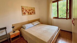 A bedroom with a bed, a nightstand, a lamp, a window with curtains, and a painting on the wall.