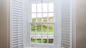 Una ventana de esquina con contraventanas blancas, vistas a colinas verdes y un muro de piedra.
