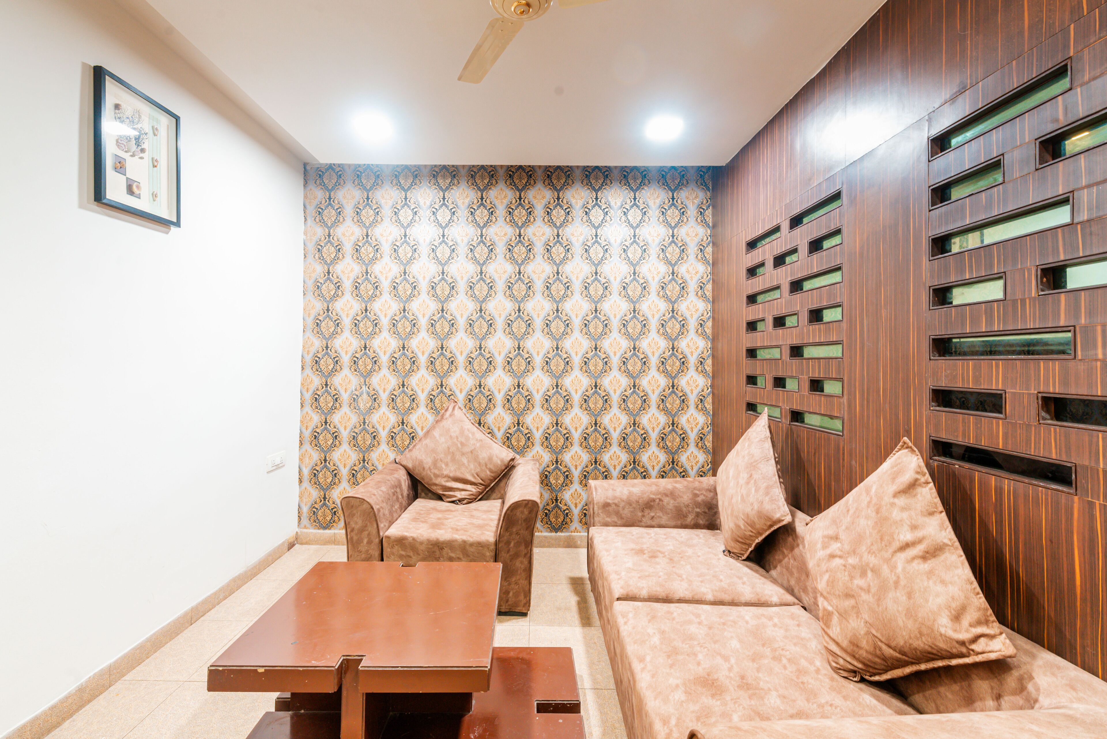 Un salón con papel tapiz estampado, un ventilador de techo, una pared de madera con ventanas y un conjunto de sofás.