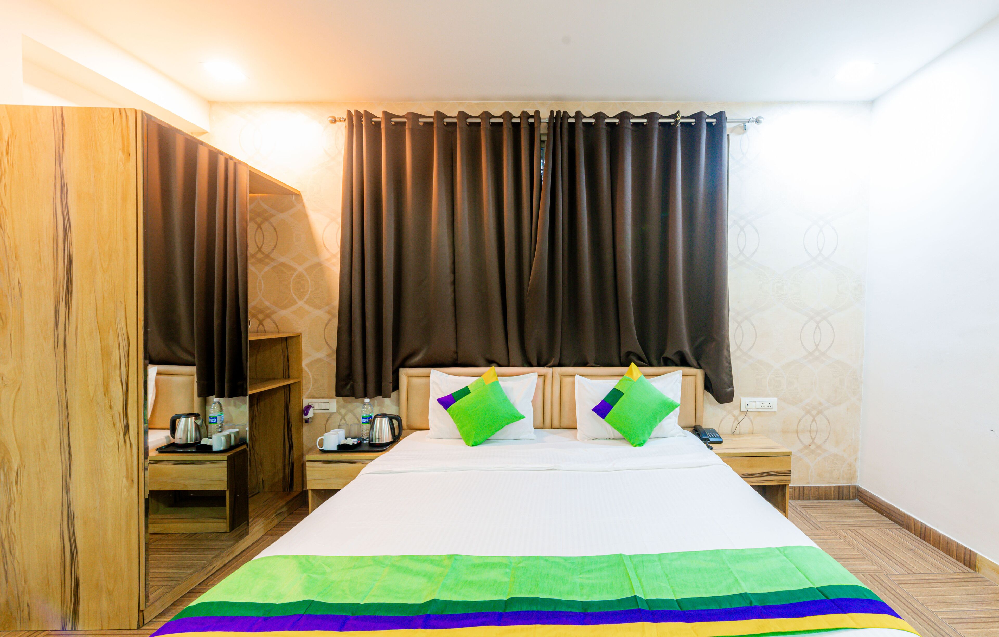 A hotel room with a large bed, a wooden wardrobe, a bedside table with a kettle, and a nightstand with a phone and a bottle.
