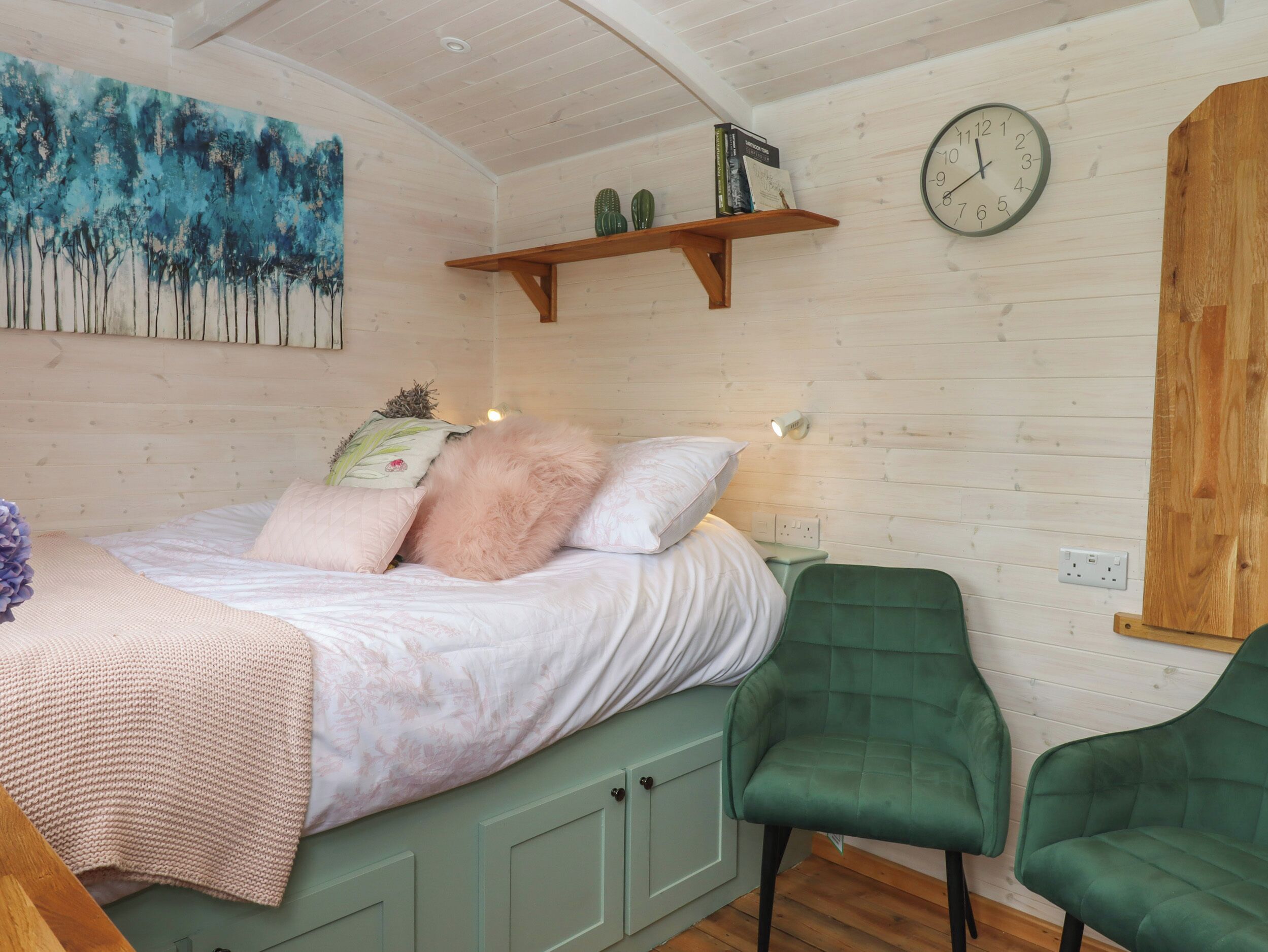 Un dormitorio acogedor con una cama, almohadas, una silla verde, un reloj y una estantería con libros y un cactus.