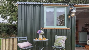 A small, green metal shed with a window and a door, a table with a vase of flowers and croissants, two chairs, and a wooden deck. - Lottie's Hut (Okehampton)