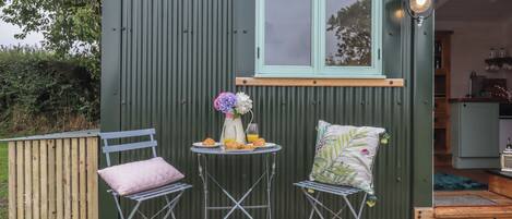 A small, green metal shed with a window and a door, a table with a vase of flowers and croissants, two chairs, and a wooden deck.