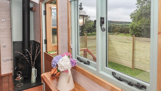 A room with a wooden bench, a vase with flowers, a lantern, and a view of a wooden deck and greenery outside. - Lottie's Hut (Okehampton)